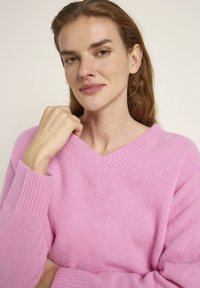 Rosa V-Ausschnitt-Pullover aus weichem Strickstoff, mit gerippten Bündchen und entspannter Passform. Die Textur wirkt flauschig und leicht.
