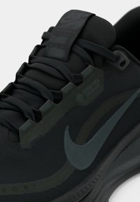 Gros plan d'une chaussure de course Nike noire montrant les lacets, l'étiquette de la languette, la marque Gore-Tex et le logo Swoosh texturé sur le côté.
