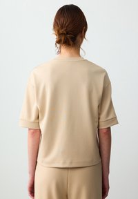 Beżowa bluzka z krótkim rękawem, z okrągłym dekoltem i podwiniętymi rękawami. Gładka faktura, luźny krój, zestawiona z dopasowanymi dolnymi elementami.
