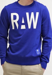 Blå sweatshirt i bomullsblandning, med stor vit "RAW" text och en liten etikett längst ner till vänster. Ribbade halsringning och ärmslut.