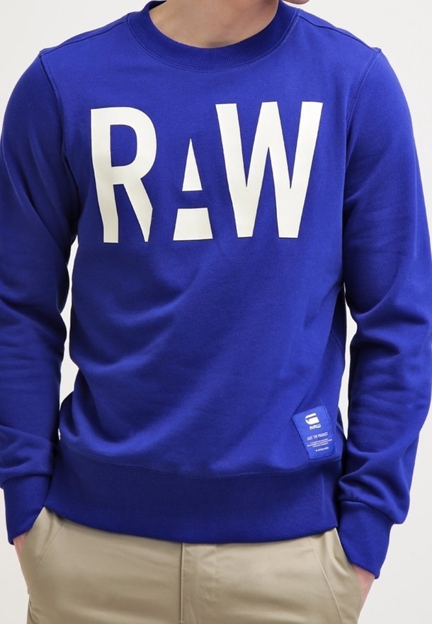 Blå sweatshirt i bomullsblandning, med stor vit "RAW" text och en liten etikett längst ner till vänster. Ribbade halsringning och ärmslut.