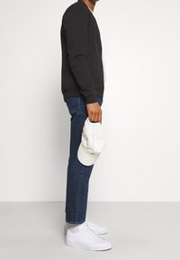 Černá mikina, bílý tričko, modré džíny a bílé tenisky. Drží bílé čepice. Hladká textura, ležérní střih a jednoduchý design.