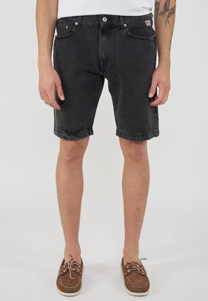 Uomo che indossa pantaloncini di jeans neri con orli risvoltati, camicia bianca e scarpe da barca marroni, in piedi contro uno sfondo chiaro e semplice.