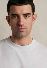 Biała bluza z okrągłym dekoltem, o miękkiej teksturze i ściągaczami na rękawach. Zbliżenie na twarz mężczyzny z krótkimi włosami i neutralnym wyrazem twarzy.