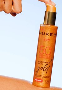NUXE Sun SPF 50 sololja i en strålande orange flaska, med en guldpump och glittrande konsistens, placerad ovanför solbrun hud. 150 ml.