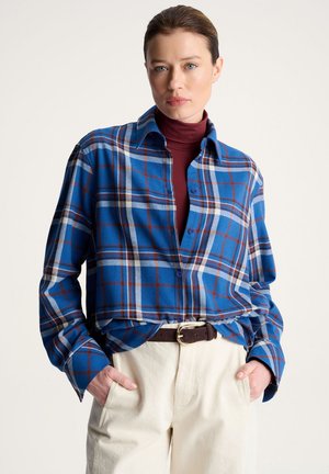 Kobieta ubrana w niebieską koszulę w kratę zapinaną na guziki, na bordowym golfie, beżowe spodnie i brązowy pasek, stoi ze rękami w kieszeniach.