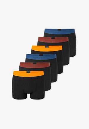 Oberkörperfreier Mann, der schwarze Boxer-Shorts mit einem orangefarbenen Bund trägt, steht vor einem schlichten hellen Hintergrund und hält die Hände vor sich zusammen.