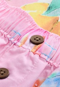 Close-up van roze stof met elastische plooien en bruine viergaatjes knopen, met een kleurrijke bedrukte achtergrond.