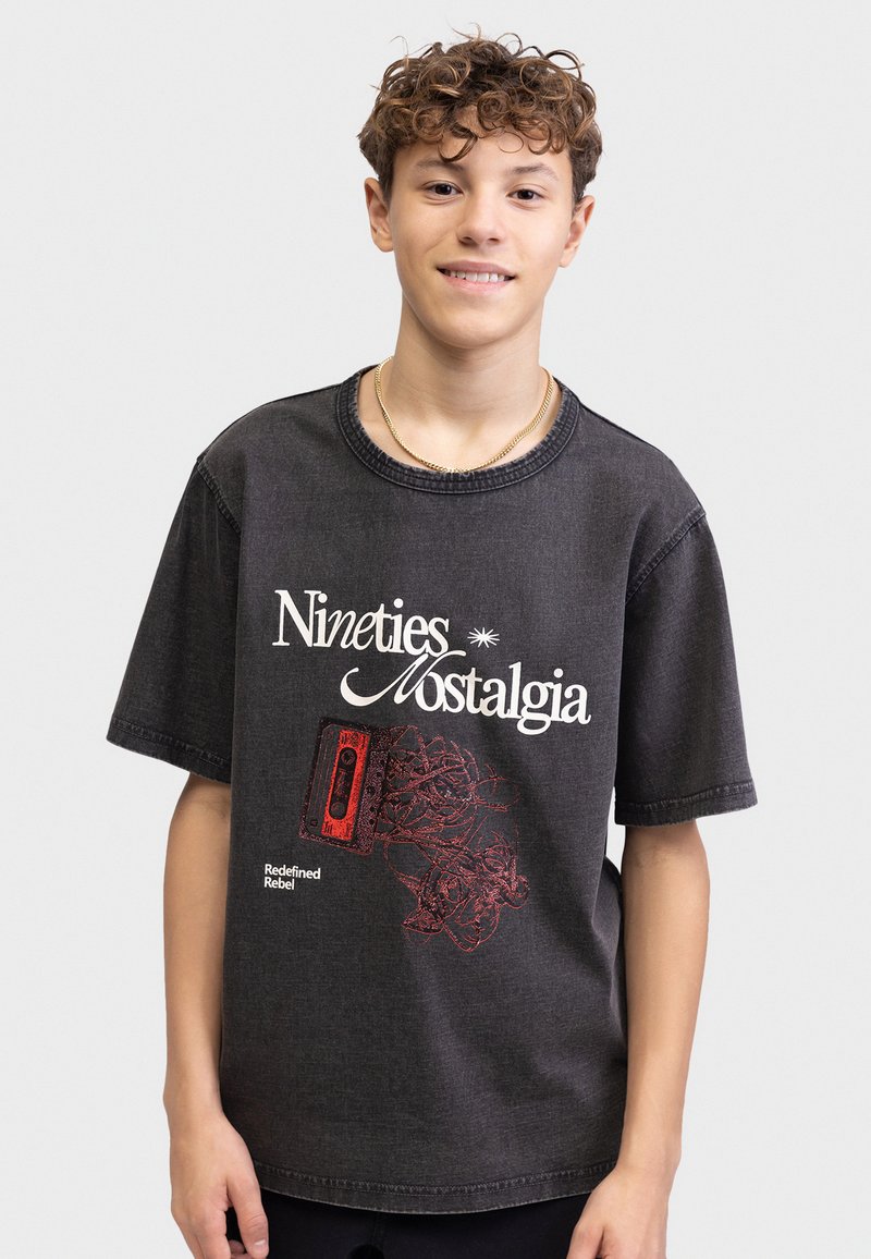 Έφηβος με σγουρά μαλλιά που φοράει μαύρο μπλουζάκι με τη φράση «Nineties Nostalgia» και γραφικά μιας κόκκινης κασέτας και μπερδεμένου μαγνητοταινίας.