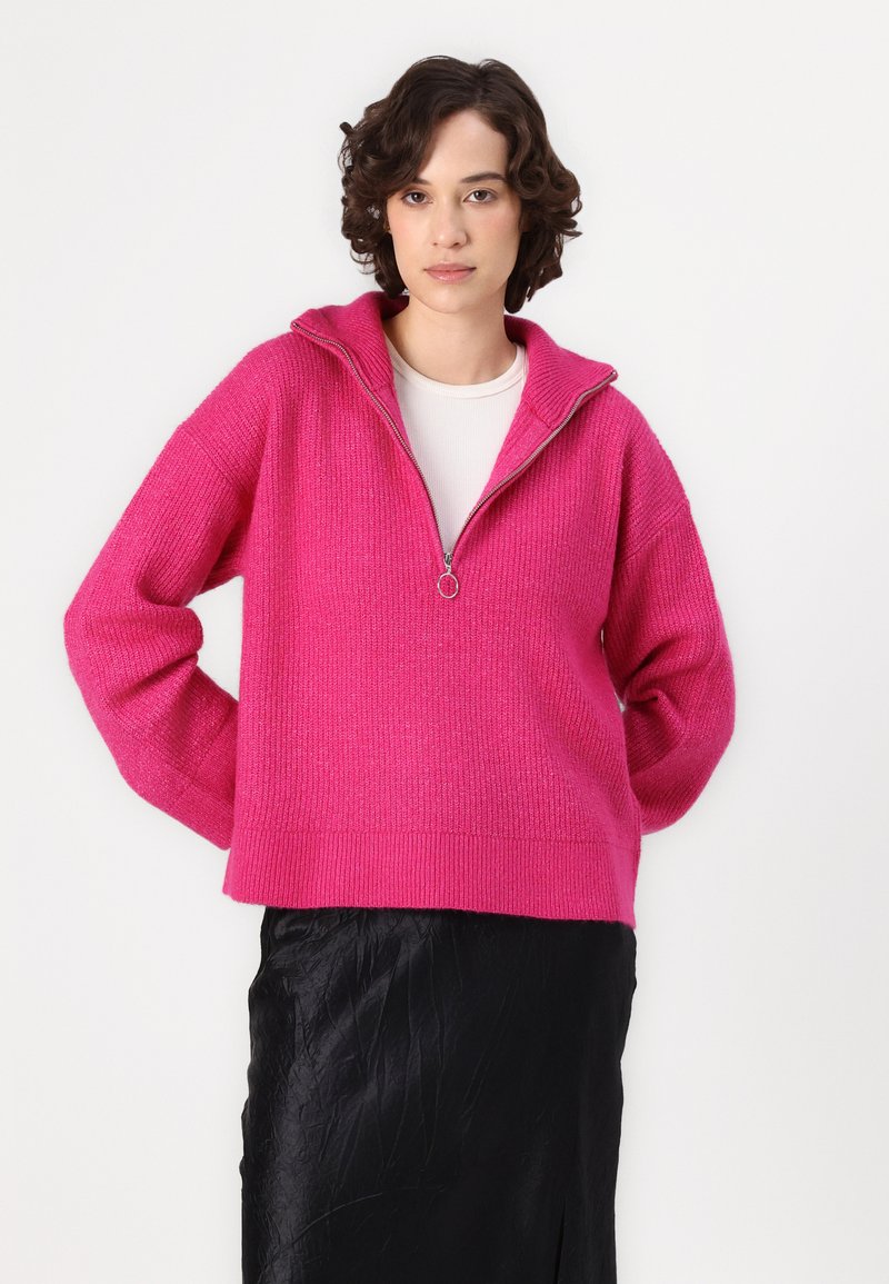 Sweter w kolorze fuksji, dziany, z ćwierć zamkiem, z prążkowanymi mankietami i dolnym wykończeniem. Noszony na białej bluzce, zestawiony z czarną teksturowaną spódnicą.