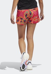 Persona caminando alejándose, vistiendo zapatillas de correr blancas, calcetines blancos, camiseta negra y pantalones cortos rojos con un patrón floral en naranja y púrpura.