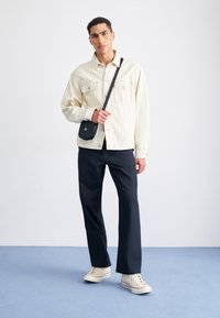 Jasno beżowa marynarka w paski z przednimi kieszeniami, czarna torebka na ramię, ciemne spodnie oraz beżowe sneakersy z kolorowymi paskami na podeszwach.