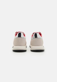 Μπεζ σουέτ αθλητικά παπούτσια με λευκές και κόκκινες λεπτομέρειες, που διαθέτουν γυαλιστερό τακούνι και μαύρη καουτσούκ σόλα. Η οπίσθια άποψη παρουσιάζει μια στρογγυλεμένη σχεδίαση.