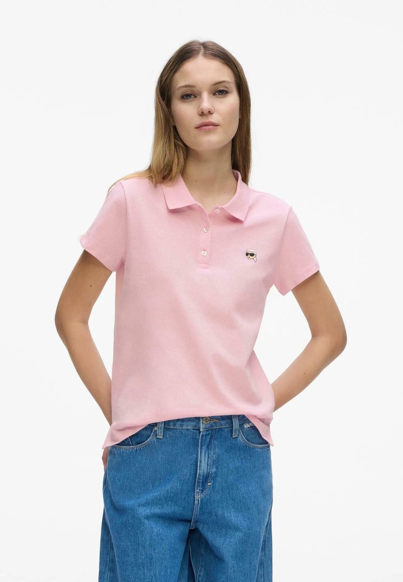 Jeune femme portant un polo rose clair à col avec un petit logo visage brodé et un jean en denim bleu, debout les mains derrière le dos.