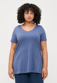 Una persona con cabello rizado está de pie contra un fondo liso, vestida con una camiseta azul de cuello en V y jeans azul oscuro, mirando directamente a la cámara.