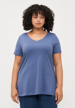 En person med krøllete hår står mot en enkel bakgrunn, iført en blå V-hals T-skjorte og mørkeblå jeans, og ser direkte inn i kameraet.