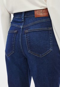 Σκούρο μπλε τζιν παντελόνια με ψηλή μέση, με πίσω τσέπες, διακριτικές λεπτομέρειες ραφής και καφέ δερμάτινη ετικέτα μάρκας στη μέση.