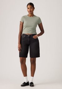 Γυναίκα που στέκεται, φοράει πράσινη ριγέ κοντή μπλούζα, μαύρα τζιν σορτς μέχρι το γόνατο, λευκές κάλτσες και μαύρα μοκασίνια, ουδέτερο φόντο.