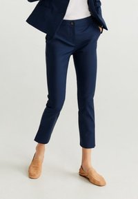Námořnicky modré kalhoty na míru s hladkou texturou, zkrácené délky, přední kapsy a zapínání na jeden knoflík. Kombinovány s béžovými nazouvacími botami.