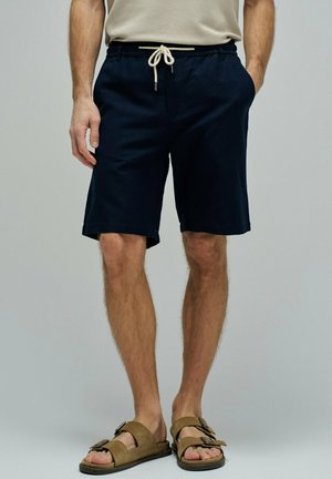 Hombre con pantalones cortos azul marino con cordón y sandalias de ante marrón con doble hebilla, de pie con una mano en el bolsillo sobre fondo gris.