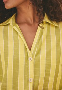 Camicia a righe verdi con colletto, chiusura a bottoni e una texture di tessuto leggero con righe bianche a contrasto.