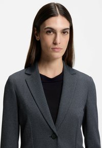Maatwerk grijze pinstripe blazer met ingeklede revers en een enkele knoopsluiting. Gdraagd over een zwart topje, met een gladde textuur en een gestructureerde pasvorm.
