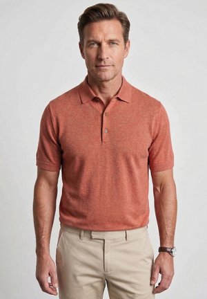 Man wearing a short-sleeve rust-colored polo shirt and beige pants, standing against a plain light background.