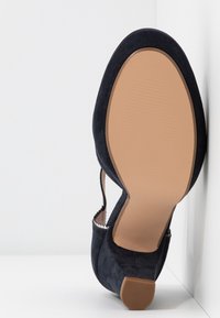 Chaussure plate pour femme en daim noir avec bout arrondi, semelle beige clair et petit talon carré, vue par le dessous sur fond blanc.