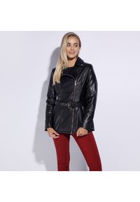 Black leather jacket with a fitted design, belted waist, asymmetric zipper, and sharp collar; paired with red pants. Smooth texture.