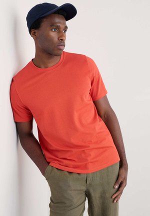 Camiseta de algodón naranja de manga corta y escote redondo, combinada con pantalones verde oliva texturizados y una gorra de béisbol azul marino.
