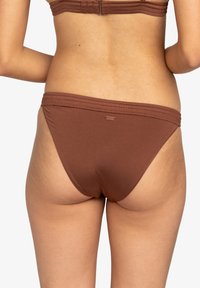 Bas de bikini marron avec un design sans coutures, en tissu extensible, et une étiquette logo discrète à l'arrière de la taille. Texture lisse et douce.