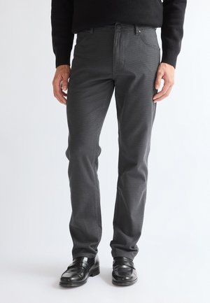 Mężczyzna noszący dopasowane, szare, w kratę spodnie, czarny sweter i czarne skórzane eleganckie buty, stojący na białym tle.