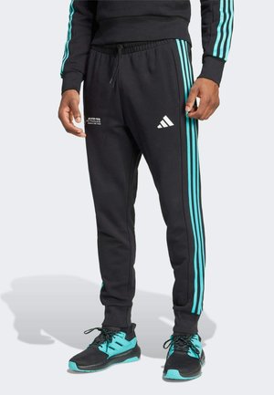 Person trägt schwarze Adidas Trainingshose mit türkisfarbenen Streifen und Logos, kombiniert mit schwarzen und türkisfarbenen Sneakers.