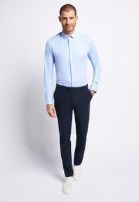 Lichtblauw overhemd met lange mouwen en een button-down kraag, gecombineerd met marineblauwe slim-fit broek en witte sneakers, allemaal gepresenteerd op een model.