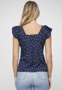 Tmavě modrá halenka s bílými srdíčkovými vzory, krátkými nabíranými rukávy a čtvercovým výstřihem na zádech. Lehký materiál, mírně přiléhavý střih.