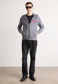 Sivi zip-up dukserica s crvenim logotipom, crna majica, tamne traperice i crne cipele. Model stoji s rukama u džepovima ispred jednostavne pozadine.