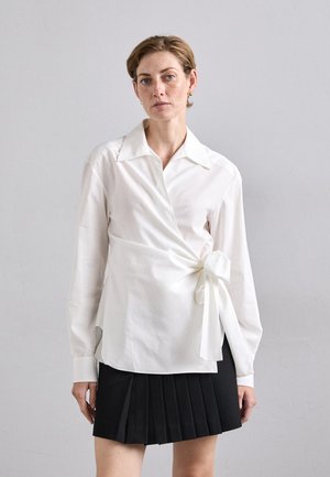 Woman with short hair wearing a white wrap blouse tied at the side and a black pleated mini skirt, standing against a plain gray background.
