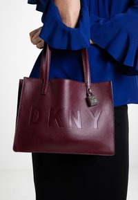 Bordowa torba DKNY wykonana z fakturowanej skóry, z wytłoczonym logo i metalowym akcentem w postaci kłódki. Szerokie uchwyty zapewniają wygodne noszenie.