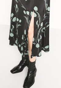 Květinové černé šaty s výškou rozparku po stehna, zdobené zelenými tulipánovými vzory. Dokončeno s černými, lesklými, úhlovými kotníkovými botami.