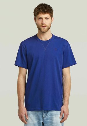T-shirt en coton bleu avec un col rond, des manches courtes et un petit logo sur la poitrine. Le tissu semble lisse et le design est décontracté.