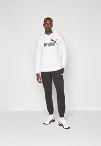 Hvid hoodie med et stort sort "PUMA" logo, parret med mørkegrå joggers med et lille logo. Model iført hvid og sorte sneakers.