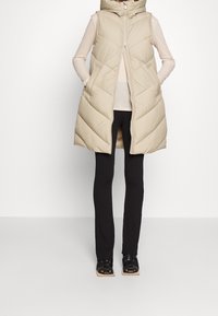 Beige gesteppte Weste mit Kapuze, Reißverschluss und seitlichen Taschen; verfügt über ein gestepptes Muster. Kombiniert mit schwarzen gerippten Hosen und Sandalen mit offenem Zehenbereich.