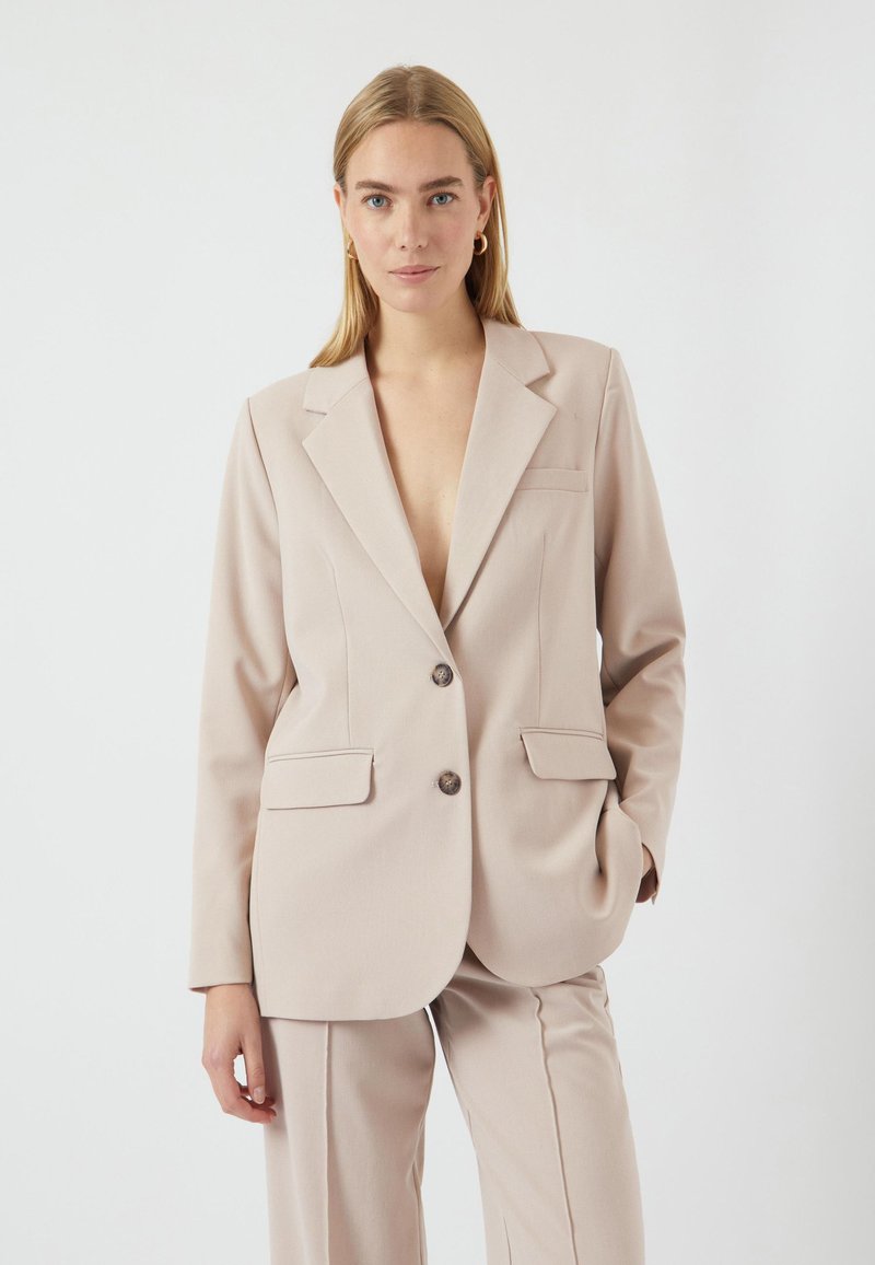 Kirkas beige muotopuku, jossa on kaksi nappia, etutaskut ja jämäkkä kaulus, yhdistettynä vastaaviin housuihin. Sileä kankaan rakenne.