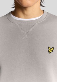 Szürke pulóver bordázott környakkal, a mellkason egy sárga és fekete hímzett madár logóval. Sima textúra, alkalmi kialakítás.