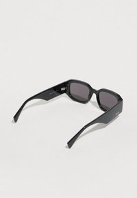 Melnas saulesbrilles ar taisnstūrveida formu, spīdīgu virsmu un tumšām lēcām. Tievas, taisnas kājiņas un smalks zīmola uzraksts vienā pusē.