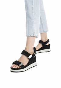 Sandali neri intrecciati con cinturini regolabili, suola a cuneo bianca a contrasto e dettagli metallici. Presentano un design open-toe e un tessuto testurizzato.