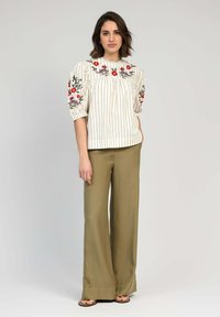 Blouse blanche à rayures avec broderie florale sur des manches bouffantes, associée à un pantalon large taille haute vert olive et des sandales plates.