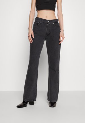 Vrouw draagt zwarte high-waisted rechte jeans, zwart cropped topje en zwarte enkellaarsjes met hak, staand tegen een effen achtergrond.