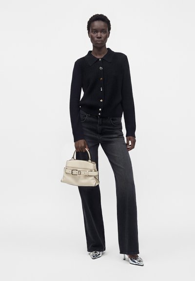 Femme portant un cardigan noir boutonné, un jean gris foncé, des talons argentés pointus, tenant un sac à main beige avec un détail ceinture sur fond blanc uni.