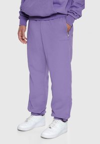 Lilla sweatpants lavet af blødt stof, med en afslappet pasform, lynlåslommer og ribbede manchetter, parret med hvide sneakers.